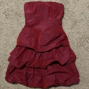 Vinröd volangklänning från Marie Blanc - Säljer en vinröd axelbandslös klänning från Marie Blanc med flera volanglager och ballongliknande kjol. Klänningen har en figurnära passform upptill och är perfekt för festliga tillfällen.