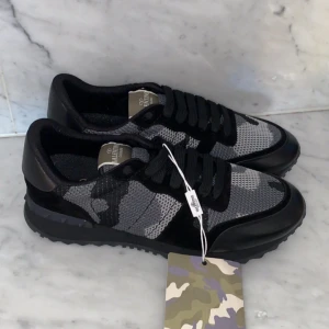 Valentino Rockrunner sneakers i svart och grått - Säljer ett par Valentino Rockrunner sneakers i svart och grått med camouflage-mönster. Skorna har svarta snören, rund tå och platt sula. Märkesdetalj på plösen och en cool, sportig look. Perfekta för dig som gillar exklusiva sneakers.