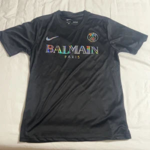 Svart PSG x Balmain fotbollströja - Snygg svart fotbollströja från Nike med Paris Saint-Germain och Balmain Paris-logga på bröstet. Tröjan har korta ärmar och är tillverkad i Dri-Fit material som andas. Perfekt för dig som gillar unika samarbeten och stilren design.