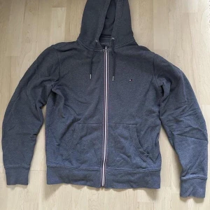 Tommy Hilfiger mörkgrå hoodie - Snygg och stilren mörkgrå zip-up hoodie. Huvan är fodrad. Fickor framtill. Dragkedjan är tvåfärgad. Perfekt för både vardag och fritid.