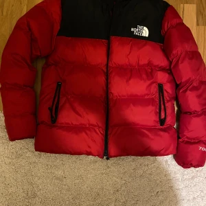 Röd pufferjacka från The North Face - Säljer en röd och svart pufferjacka från The North Face i storlek M. Jackan har lång ärm, dragkedja framtill och två fickor med dragkedja. Perfekt för kyliga dagar och har klassisk logga på bröstet och ryggen.