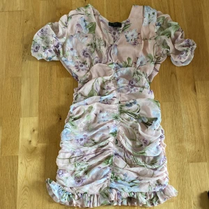 Blommig kortklänning från AX Paris - Supersöt blommig kortklänning från AX Paris i ljusrosa med gröna och lila blommor. Klänningen har puffiga korta ärmar, v-ringning, volangkant nertill och rynkade detaljer framtill. Perfekt för sommarens alla tillfällen!