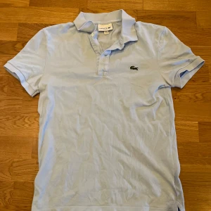 Lacoste - Säljer en klassisk ljusblå piké t-shirt från Lacoste med korta ärmar och den ikoniska krokodilloggan på bröstet. Perfekt för en clean och sportig stil. Krage och knappar framtill. Hör av dig vid frågor.