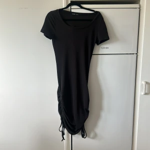 Svart kortärmad klänning med snörning från SHEIN - Svart ribbad klänning från SHEIN med korta ärmar och rund halsringning. Klänningen har snörningsdetalj på sidan som ger en snygg drapering och gör längden justerbar. Perfekt för dig som gillar en enkel men trendig stil.