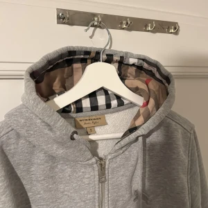 Grå hoodie från Burberry med rutigt foder - Säljer en grå hoodie från Burberry med dragkedja och klassiskt rutigt foder i huvan. Tröjan har justerbar huva och mjukt material på insidan. Perfekt för en stilren och bekväm look.
