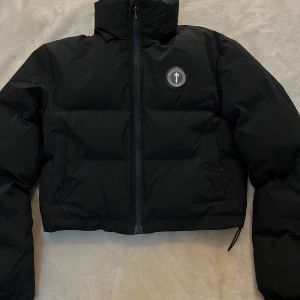 Svart pufferjacka från Trapstar - Säljer en svart pufferjacka från Trapstar med avtagbar huva och dragkedja framtill. Jackan har logotyp på bröstet och stor Trapstar-text broderad på ryggen. Perfekt för kyliga dagar och riktigt snygg streetstil.