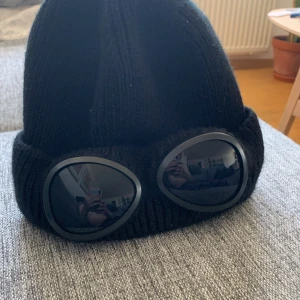 Svart stickad mössa med goggles - Cool svart stickad mössa med inbyggda goggles framtill. Perfekt accessoar för att sticka ut och hålla värmen under kyliga dagar. Mössan har en ribbad struktur och är helsvart.