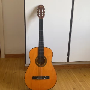 Klassisk gitarr från Kayok  - Säljer en klassisk akustisk gitarr från Kayok i trä med naturlig ljusbrun färg på framsidan och mörkbrun baksida. Gitarren har sex nylonsträngar och traditionell rosett runt ljudhålet. Perfekt för nybörjare eller som extragitarr.