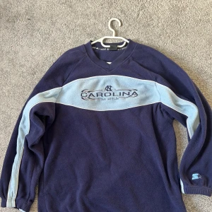 Blå långärmad tröja från North Carolina Tar Heels - Mörkblå långärmad tröja med ljusblå ränder längs ärmar och bröst. Broderad text 'Carolina Tar Heels' framtill. V-ringad hals och sportig design, perfekt för dig som gillar college-stil.