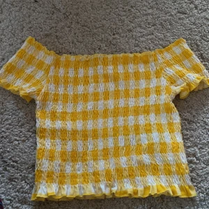 Gul och vit smockad topp från FB Sister - Säljer en söt smockad topp från FB Sister i gult och vitt rutigt mönster. Toppen har korta ärmar och är croppad med volangkant nertill. Perfekt för varma dagar!