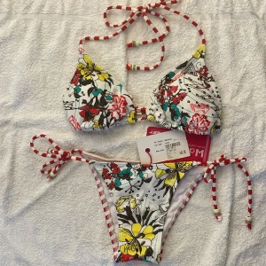 Färgglad triangel bikini från Protest med blommigt och randigt mönster - Säljer en snygg triangel bikini från Protest med blommigt mönster på ena sidan och rödvita ränder på andra. Överdelen har knyt i nacken och ryggen, och underdelen har knyt i sidorna. Färgerna är vit, röd, gul och inslag av svart och grön. Bikinin är helt oanvänd och har trosskydd och prislappar kvar! Pris kan diskuteras.