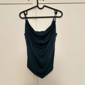 Mörkgrön satin linnetopp - Snygg mörkgrön linnetopp i satin med draperad front och tunna axelband. Perfekt att styla till jeans eller kjol för en trendig look.