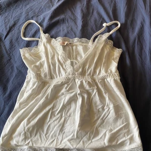 Vit spetsdetaljerad linnetopp - Söt vit linnetopp med tunna axelband och spetsdetaljer längs urringning och nederkant. Perfekt för varma dagar eller som ett lager under andra plagg. Mjuk och lätt i materialet.