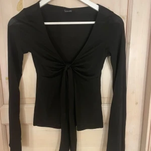 Svart långärmad topp med knyt från Gina Tricot - Säljer en svart långärmad topp från Gina Tricot i storlek XXS. Men den passar även mig som i valpiga fall har S. Toppen har djup v-ringning och knyt framtill, vilket ger en snygg och trendig look. Perfekt att styla med jeans eller kjol.