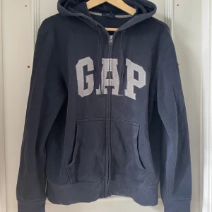 Mörkblå hoodie med dragkedja från GAP - Mörkblå hoodie från GAP med dragkedja framtill och stora vita bokstäver på bröstet. Tröjan har huva och två fickor framtill. Perfekt för en avslappnad stil, så najs till sommaren med ett par ljusblå shorts/jeans och vitt linne under till exempel 💕