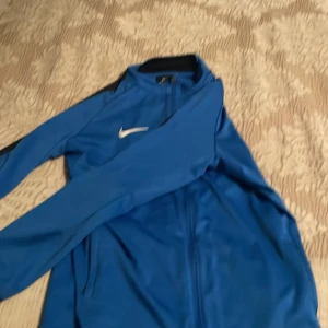 Blå träningsjacka från Nike Dri-Fit - Säljer en blå Nike Dri-Fit träningsjacka med dragkedja och lång ärm. Jackan har svart detalj på axlarna och klassisk vit Nike-logga på bröstet. Perfekt för fotboll eller träning, tillverkad i lätt syntetmaterial.