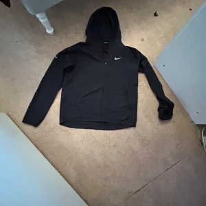 Svart hoodiejacka från Nike - Svart hoodiejacka från Nike med dragkedja och huva. Jackan har lång ärm och en liten vit Nike-logga på bröstet. Perfekt för en sportig stil och enkel att matcha med det mesta.