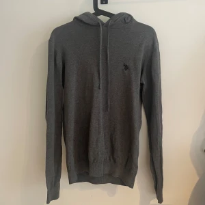 Grå stickad hoodie från U.S Polo assn. - Säljer en stilren grå hoodie från U.S Polo assn med liten broderad logga på bröstet. Tröjan har långa ärmar, huva med dragsko och ribbade muddar. Perfekt för en avslappnad och snygg look. 