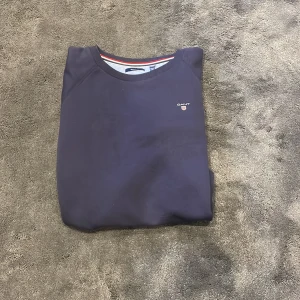 Mörkblå sweatshirt från GANT - Säljer en stilren mörkblå sweatshirt från GANT med rund halsringning och diskret logga broderad på bröstet. Tröjan har långa ärmar och klassisk passform. Perfekt för en avslappnad och snygg look. (Storlek 170 cm)