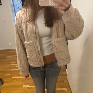 Beige quiltad jacka - Säljer en beige quiltad jacka med lång ärm och två stora fickor framtill. Jackan har en rak passform och stängs med dragkedja. Perfekt för övergångssäsonger och enkel att matcha till olika outfits.