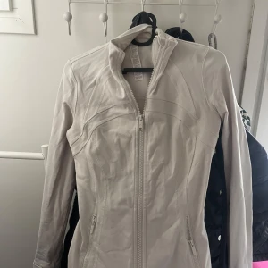 Vit jacka från Lululemon med dragkedja - Säljer en vit jacka från Lululemon i storlek 4 (XS). Jackan har figurnära passform, hel dragkedja framtill och två fickor med dragkedja. Tillverkad i mjukt och stretchigt material, perfekt för träning eller vardag. Dragkedjan saknar en hårögla. Köpt begagnat.🩷