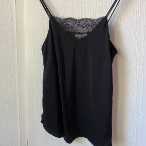 Svart linnetopp med spetsdetalj - Säljer en svart linnetopp med tunna axelband och v-ringning. Toppen har fina spetsdetaljer vid halsringningen och är tillverkad i ett mjukt, glansigt material. 