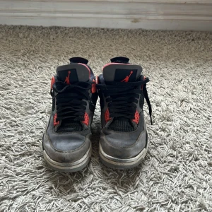 Nike Air Jordan 4 Retro svart/grå/röd - Säljer ett par Nike Air Jordan 4 Retro sneakers i svart, grått och rött. Skorna har svarta snören, röda detaljer och vit sula. Klassisk Jumpman-logga på hälen och tungans framsida. Perfekta för dig som gillar streetwear och basketstil.