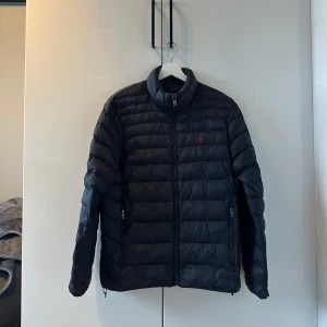 Blå dunjacka från Polo Ralph Lauren - Blå dunjacka från Polo Ralph Lauren med klassisk quiltad design och diskret röd logga på bröstet. Jackan har hög krage, dragkedja framtill och två sidofickor med dragkedja. Perfekt för kyliga dagar och enkel att matcha med allt. Nypris 4500 mitt pris 1999. Pris kan diskuteras! Skriv vid minsta fråga! 