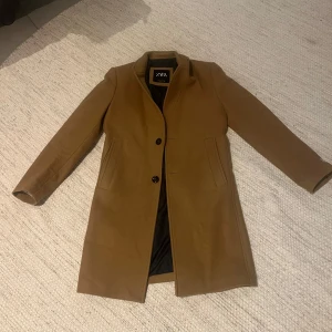 Klassisk brun kappa från Zara - Snygg brun kappa från Zara i stilren modell med två knappar framtill och raka fickor. Jackan har lång ärm och är fodrad med svart tyg på insidan. Perfekt för dig som gillar en clean och tidlös look. Kappan kostade egentligen 1200 kr men säljs för 299 kr, och har bara använts få gånger.