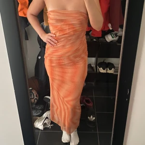 Orange mönstrad långklänning - Säljer en snygg orange långklänning med coolt vågigt mönster. Klänningen är axelbandslös och har en figurnära passform som framhäver siluetten. Materialet känns lätt och stretchigt, perfekt för dig som vill sticka ut med färg och form. Dragkedja i ryggen för enkel påklädning.