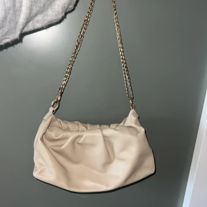 Beige axelväska med kedja - Snygg beige axelväska med mjukt, rynkat skinnmaterial och guldfärgad kedja som axelrem. Perfekt storlek för det viktigaste och ger en cool vibe till din outfit. Enkel design med lyxig känsla tack vare kedjan.