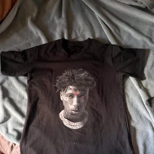 Svart nba youngboy t-shirt  - Svart t-shirt från Vlone med stor röd V-logga på ryggen och tryck av nba youngboy på framsidan. Klassisk rund hals och korta ärmar. Materialet är mjuk bomull och passformen är normal. Perfekt för dig som gillar streetwear och vill sticka ut.