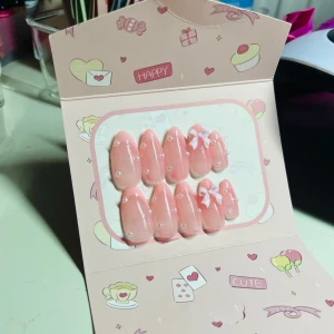 Söta rosa lösnaglar med pärlor och rosetter - Ett set med 10 lösnaglar i en mjuk rosa färg, dekorerade med små pärlor och två vita rosetter. Naglarna har en elegant, mandelformad design och kommer med tillbehör som lim, nagelfil, träpinne och alkoholpad för enkel applicering. Perfekt för dig som vill ha gulliga och trendiga naglar.