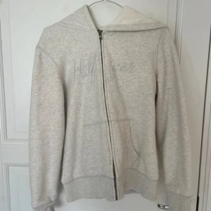 Ljusgrå hoodie från Tommy Hilfiger - Mysig ljusgrå hoodie från Tommy Hilfiger med broderad logga på bröstet. Tröjan har huva, dragkedja och två fickor framtill. Insidan är fodrad med mjuk fleece som gör den extra skön. Perfekt för kyliga dagar och chill outfits.