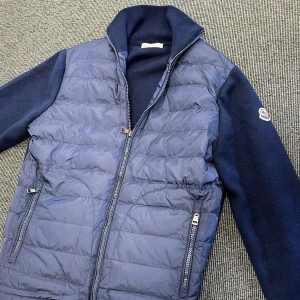 Blå cardigan från Moncler - Snygg marinblå pufferjacka från Moncler med stickade ärmar och quiltad front. Jackan har dragkedja framtill, två fickor med dragkedja och klassisk Moncler-logga på ärmen. Perfekt för dig som vill ha en stilren och sportig look.