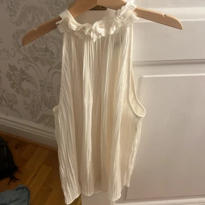 Vit plisserad blus med blommor Zara - Superfin vit plisserad blus från Zara med ärmlös design och hög halsringning dekorerad med 3D-blommor. Blusen har en lätt och flowy känsla, knäpps i nacken och är perfekt för dig som gillar romantiska detaljer och stilren look.