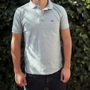 Grå Lacoste pikétröja i bomull - Snygg grå pikétröja från Lacoste med klassisk krage och två knappar framtill. Ikoniska krokodilloggan på bröstet. Tillverkad i mjuk bomull som andas. Perfekt för dig som gillar stilren och enkel design.                                                                                                                                                                              Nypris: 1209kr GORILLAPRIZE:550kr            Skick:8/10 mycket bra skick         KVITTO MEDKOMMER     