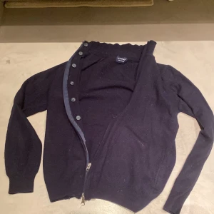 Svart kofta i kashmir/ull från Sirmoney - Mysig svart kofta från Sirmoney i en lyxig mix av kashmir och ull. Koftan har långa ärmar, knappar framtill och dragkedja nertill för extra stilpoäng. Perfekt för lager-på-lager och chill höst- eller vinterlook.