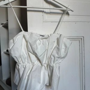 Vit omlott linnetopp från Zara - Säljer en vit linnetopp från Zara med tunna axelband och snygga omlottdetaljer framtill. Toppen har en luftig och lite voluminös form med rynk vid midjan som ger en cool siluett. Perfekt för dig som gillar stilrena och trendiga plagg.