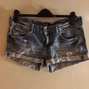Blå jeansshorts från H&M Divided - Snygga blå jeansshorts från H&M Divided med slitna detaljer och upprullade benslut. Klassisk femficksmodell med knapp och dragkedja framtill. Vet inte riktigt storlek men mellan S och M.