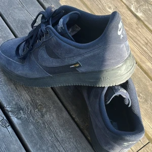 Mörkblå Nike Air Force 1 sneakers - Snygga mörkblå Nike Air Force 1 sneakers i mocka och textil med klassisk siluett och svart sula. Skorna har snörning och diskret Cordura-detalj på sidan. Perfekta för dig som gillar streetstyle och vill ha en ikonisk sneaker i garderoben.