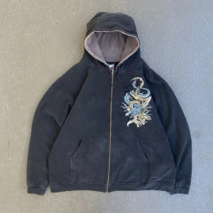 Svart hoodie med drakbrodyr Top Heavy - Svart hoodie från Top Heavy i storlek XXL med fet broderad drake i blått och beige på både fram- och baksida. Tröjan har dragkedja, huva med ljus insida och kängurufickor. Materialet är 80% bomull och 20% polyester. Perfekt för dig som gillar streetwear vibes.