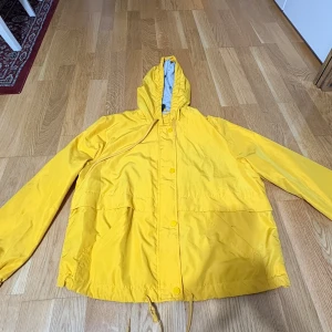 Gul vindjacka med huva och snörning - Snygg gul vindjacka med huva och dragsko i midjan. Jackan har långa ärmar, tryckknappar framtill och stora fickor. Perfekt för blåsiga dagar när du vill sticka ut lite extra. Enkel och clean design som passar till det mesta.