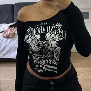 Svart offshoulder croppad topp med tryck  - Säljer en svart croppad topp med offshoulder-design och lång ärm. Trycket framtill har en cool motorcykel, vingar och text i vitt. Toppens passform är tight 