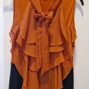 Orange och svart klänning med volanger från GEMS - Snygg kortklänning från GEMS i orange och svart. Klänningen är ärmlös med flera lager volanger framtill och en stor rosett vid halsen. Materialet är lätt och känns som polyester, perfekt för dig som gillar att sticka ut. Rak passform och färgblockad design gör den unik. 