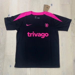 Chelsea svart och rosa fotbollströja Nike - Chelsea fotbollströja från Nike i svart med starka rosa detaljer på axlar och tryck. Kortärmad modell med rund hals och klubbmärke på bröstet. Materialet är lätt och andas, perfekt för träning eller match. Trivago-logga framtill och snygg sportig design.