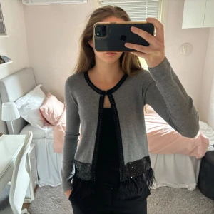 Kofta med fjädrar  - Grå stickad kofta med svarta fjädrar och små paljetter. Står storlek s/m på lappen. Man kan knäppa den vid halsen om man vill.  Skriv om du har några frågor🩷