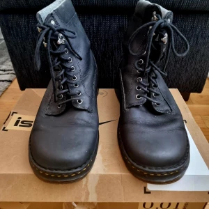 Svarta Dr. Martens boots i skinn - Klassiska svarta Dr. Martens boots i skinn med grov sula och ikoniska gula sömmar. Skorna har snörning med metallhål och hakar upptill, samt rund tå och dragflik baktill med logga. Perfekta för dig som vill ha en edgy och tidlös stil.Endast använda 2 gånger!