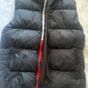 Svart dunväst från Tommy Hilfiger - Svart dunväst från Tommy Hilfiger med quiltad design och två dragkedjeförsedda fickor. Västen har en diskret logga på bröstet och är fodrad med tekniska fibrer för extra värme. Perfekt att slänga över en hoodie när det är kyligt ute. Pris går att diskuteras, den är L i barn storlek o passar bra som s/36