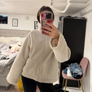 Vit teddyjacka med dragkedja - Mysig vit teddyjacka med breda ärmar och hög krage. Jackan har två stora fickor framtill och stängs med dragkedja. Perfekt för dig som gillar en chill och oversized look. Materialet är fluffigt och ger en skön vibe under kyliga dagar. Finns lite smink ovantill 